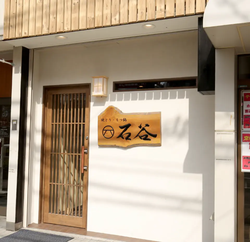 白ベースの壁に、木の「石谷」と書かれた看板がかけられていて、その隣に木製のドアがある